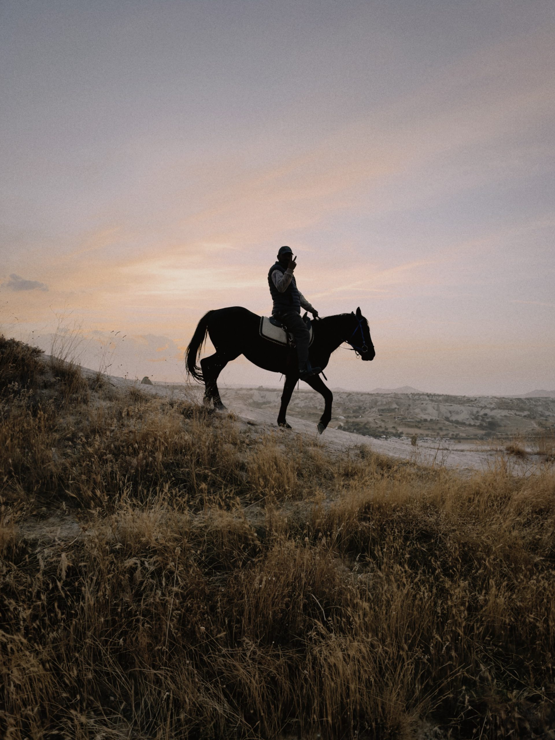 Man riding a black horse through dry grassland at sunset